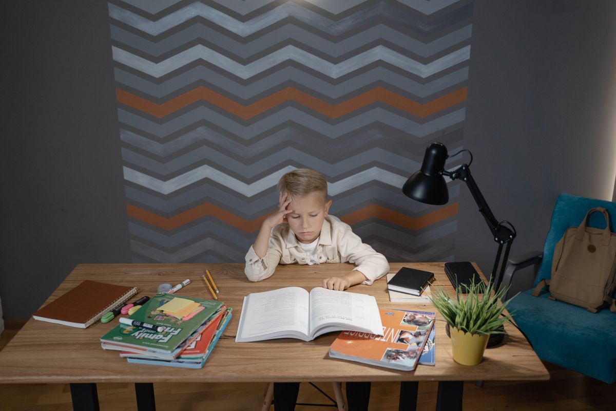 Child studying at a desk with books, a lamp, and a school bag nearby